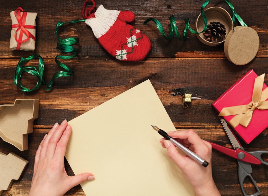A hand poses with a pen to write on a blank piece of paper. On a table surrounded by Christmas wrapping materials.