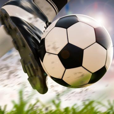 Close up of soccer player's foot kicking a soccer ball on green grass in front of a gray sky.