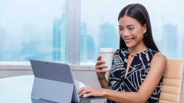 seated young female professional smiling while typing at her computer with her left hand and holding her coffee in the right hand