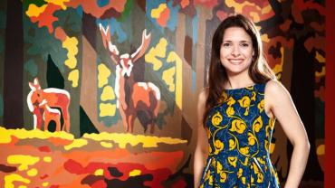 profile photo of smiling Sarah Feingold standing in front of an oil painting of family of deer