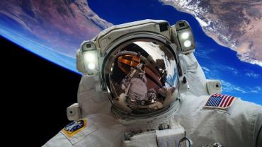 profile of an astronaut in space with the earth in the background. The astronauts face shield reflects the space station.