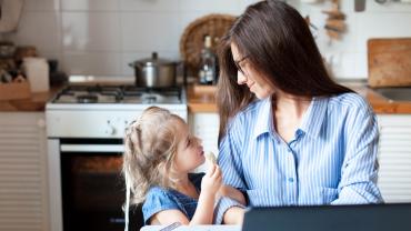  Smiling mother working on a laptop in a kitchen and her young daughter look to each other