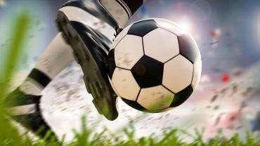 Close up of soccer player's foot kicking a soccer ball on green grass in front of a gray sky.