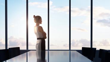 Businesswoman standing in front of a board room table looking out of a glass window in a modern office.