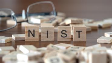Scrabble pieces on a table. Four of them are stacked next to each other, spelling out "NIST."