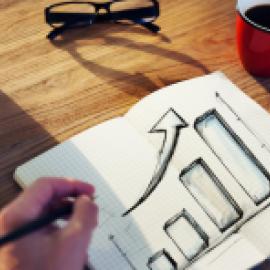 a lefthanded person draws a bar chart and a large arrow trending up in a notebook, there is also a pair of glasses, a small plant, and a cup of coffee on the table