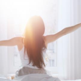 Back view of woman stretching her arms outward in the morning as she wakes up in front of white curtains and the sunlight pouring in.